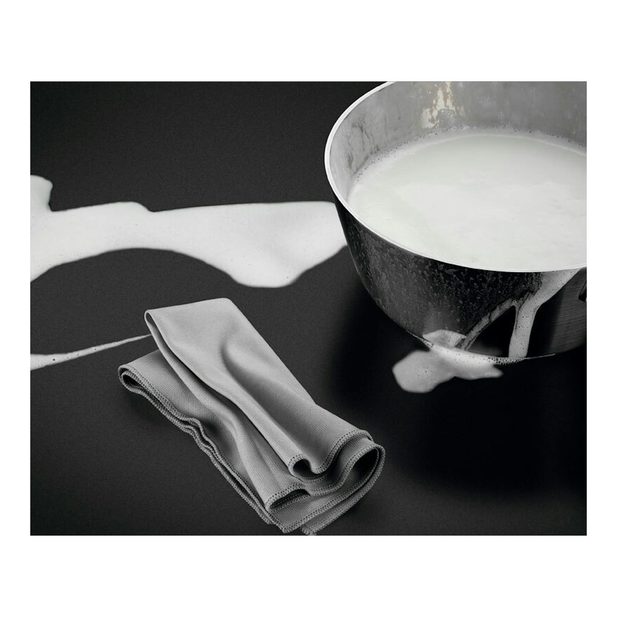 Placa de inducción AEG negra con leche derramada, paño gris y cazo metálico, vista cenital.