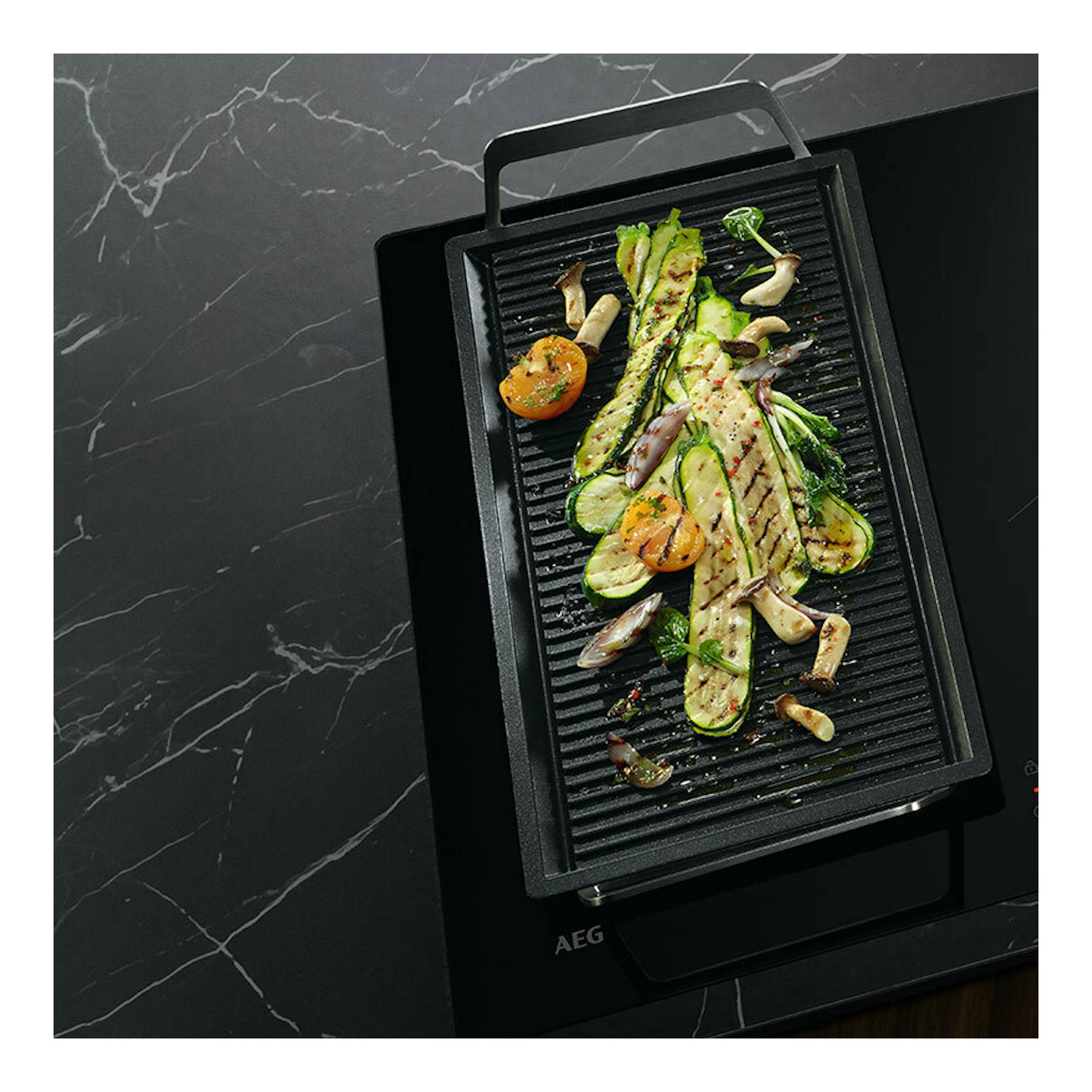 Placa de inducción AEG TO74IB00FB negra, vista cenital en ángulo, con plancha grill y verduras.