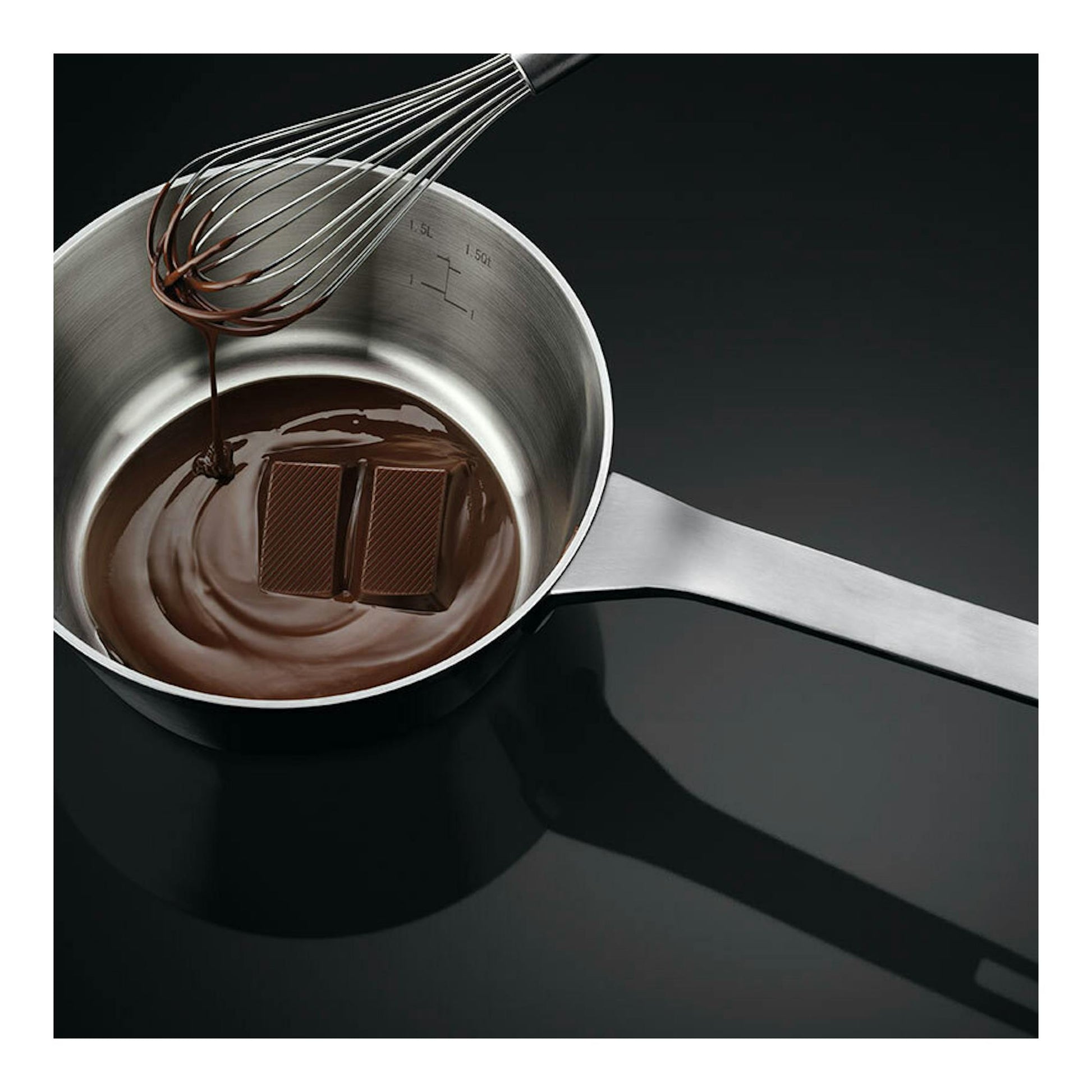 Placa de inducción negra con cazo de acero, chocolate fundido y varillas, vista superior.