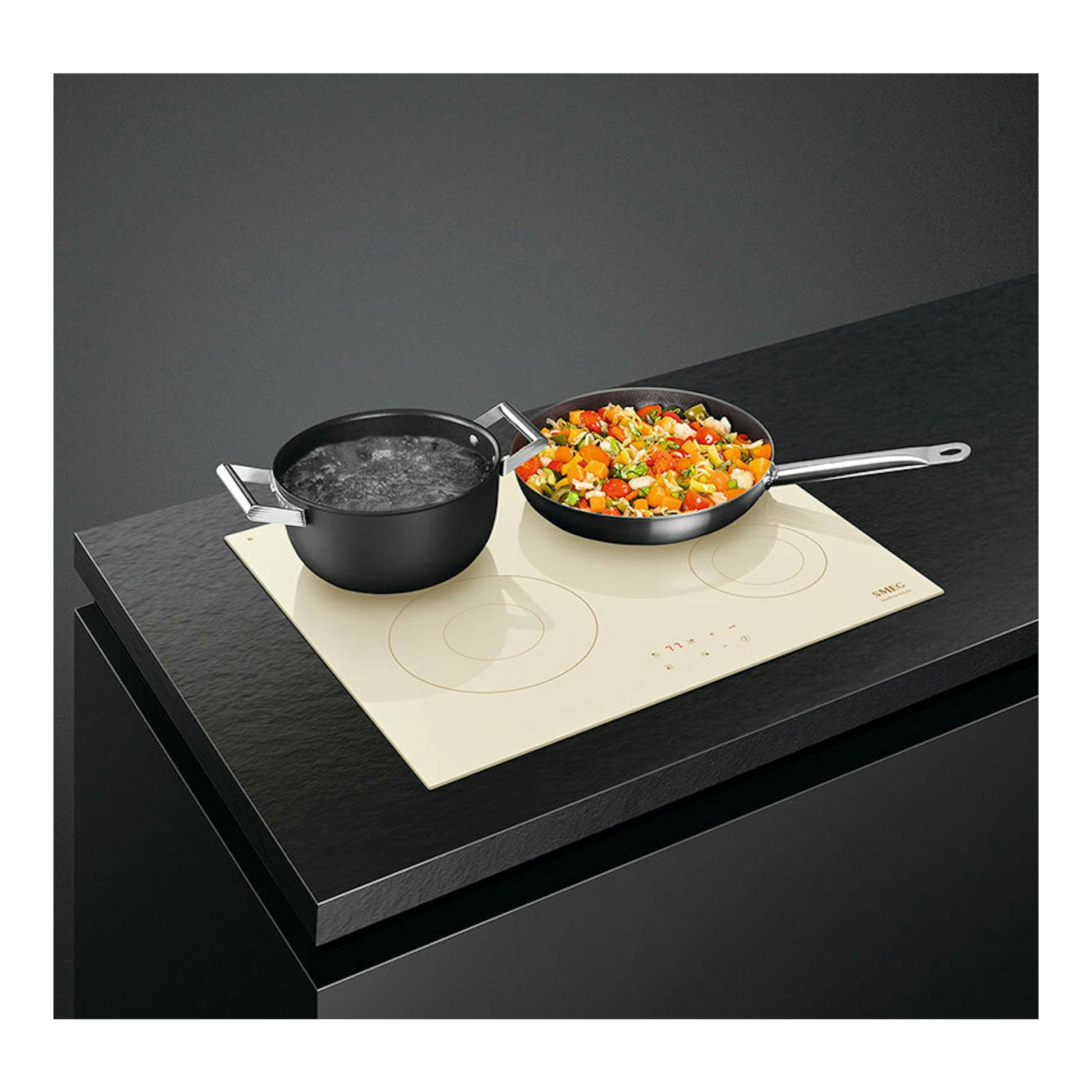 Placa de inducción Smeg SI2641DP color crema, vista en ángulo, con dos ollas sobre encimera oscura.