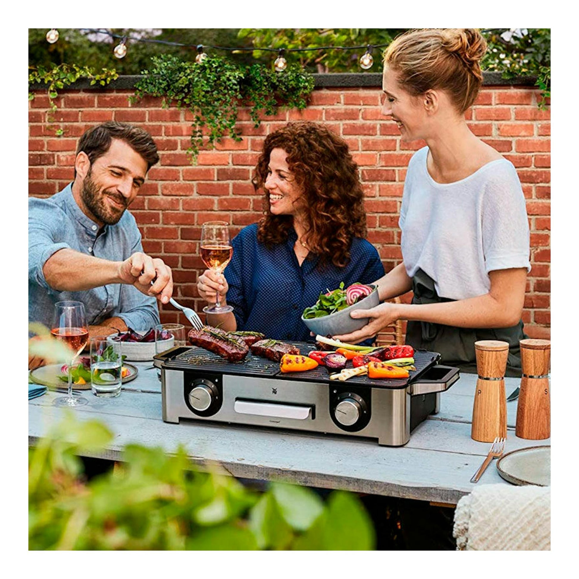 Plancha grill eléctrica WMF Master Grill LONO, vista frontal sobre una mesa, con comida a la parrilla al aire libre.