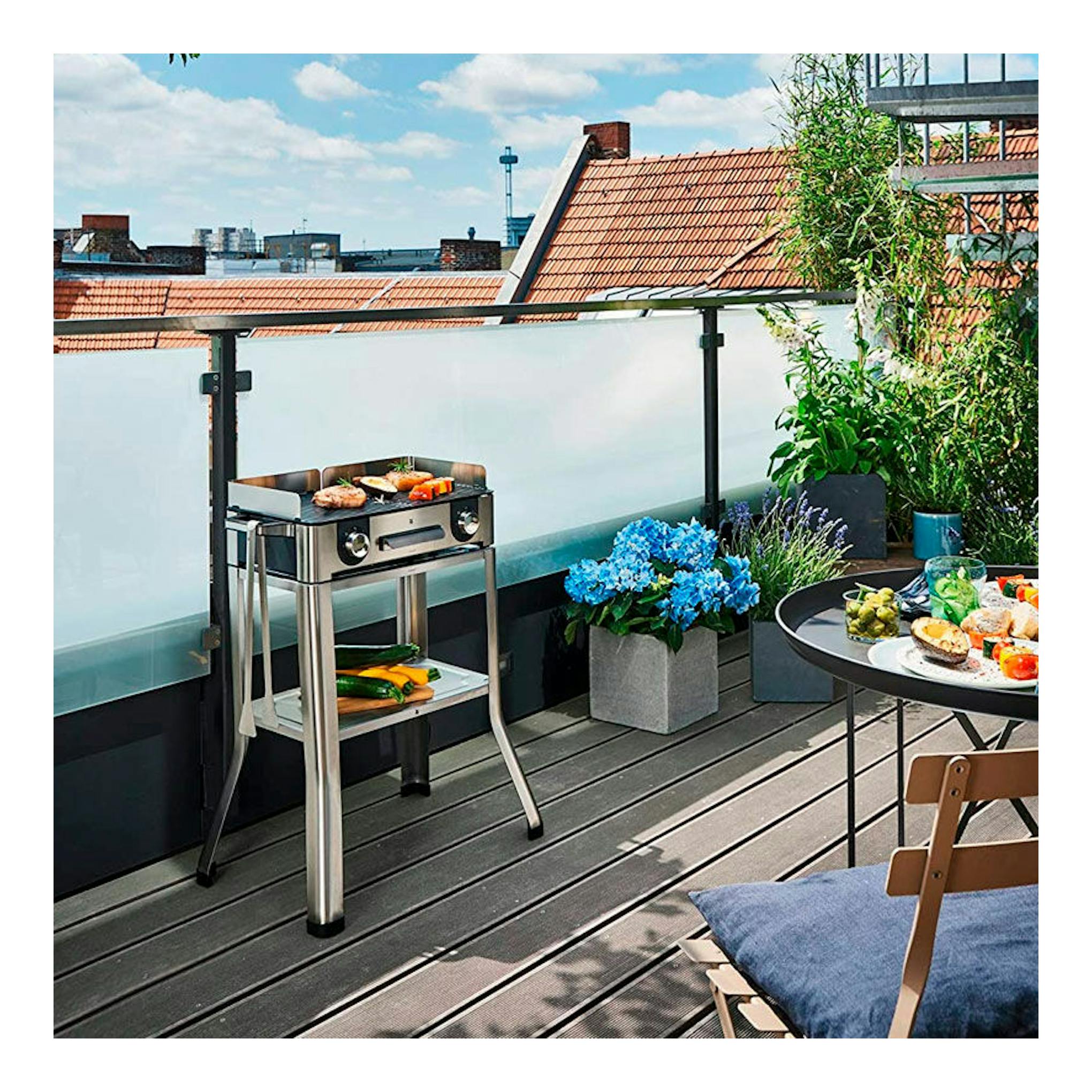 Plancha grill eléctrica WMF Master Grill LONO de acero inoxidable, vista frontal en una terraza con alimentos.