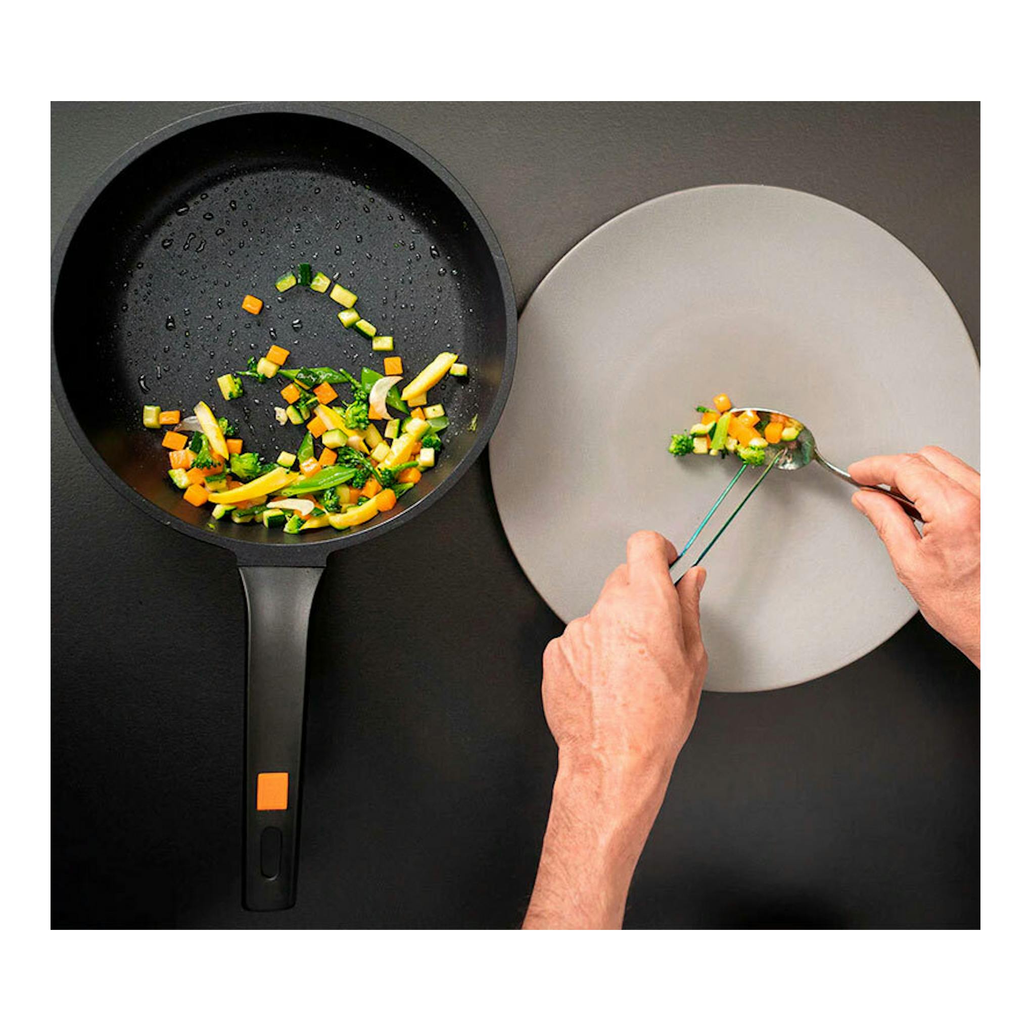 Sartén Bra Efficient negra, vista cenital, con verduras salteadas; a la derecha, manos sirviendo en plato gris.