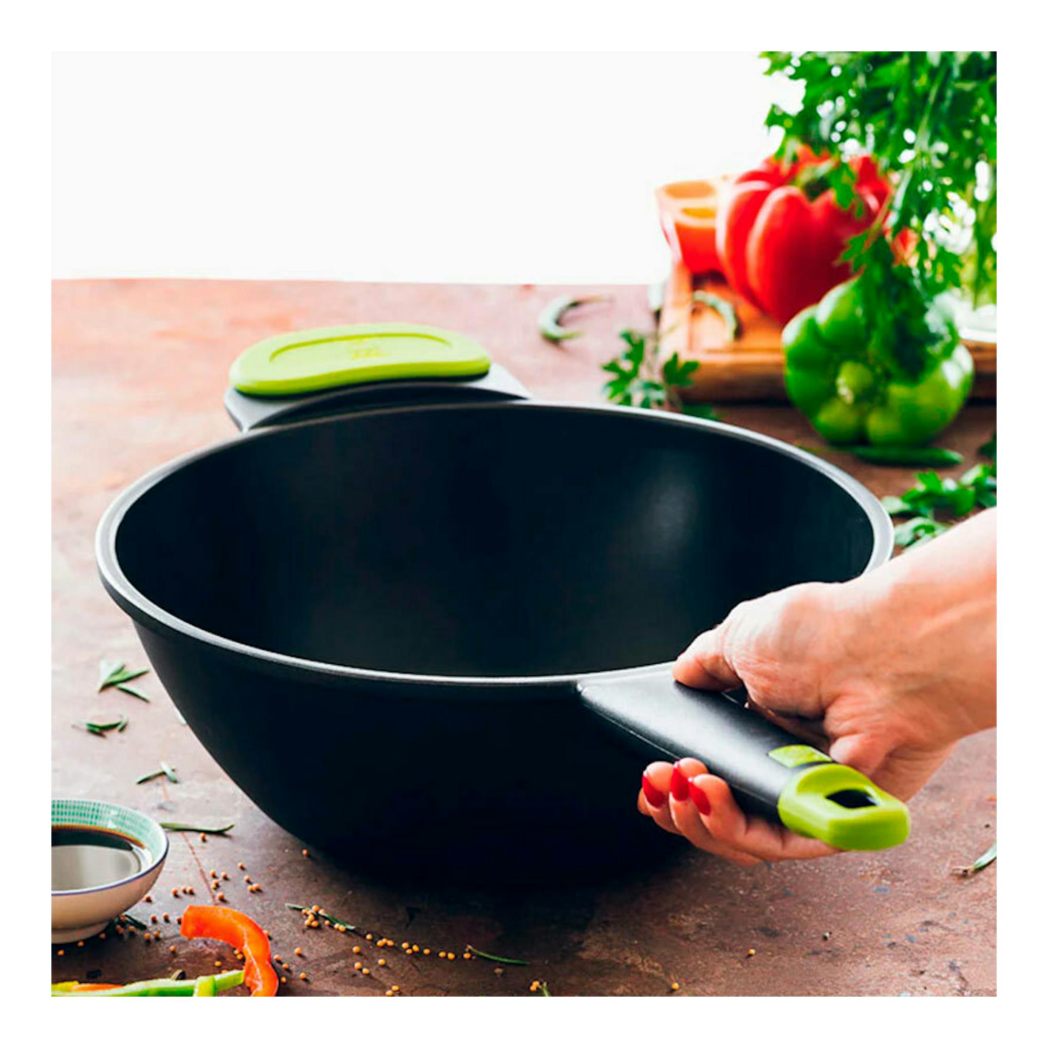 Wok negro Bra Foodie de 28 cm con mango verde, visto en ángulo sobre encimera, sostenido por una mano.