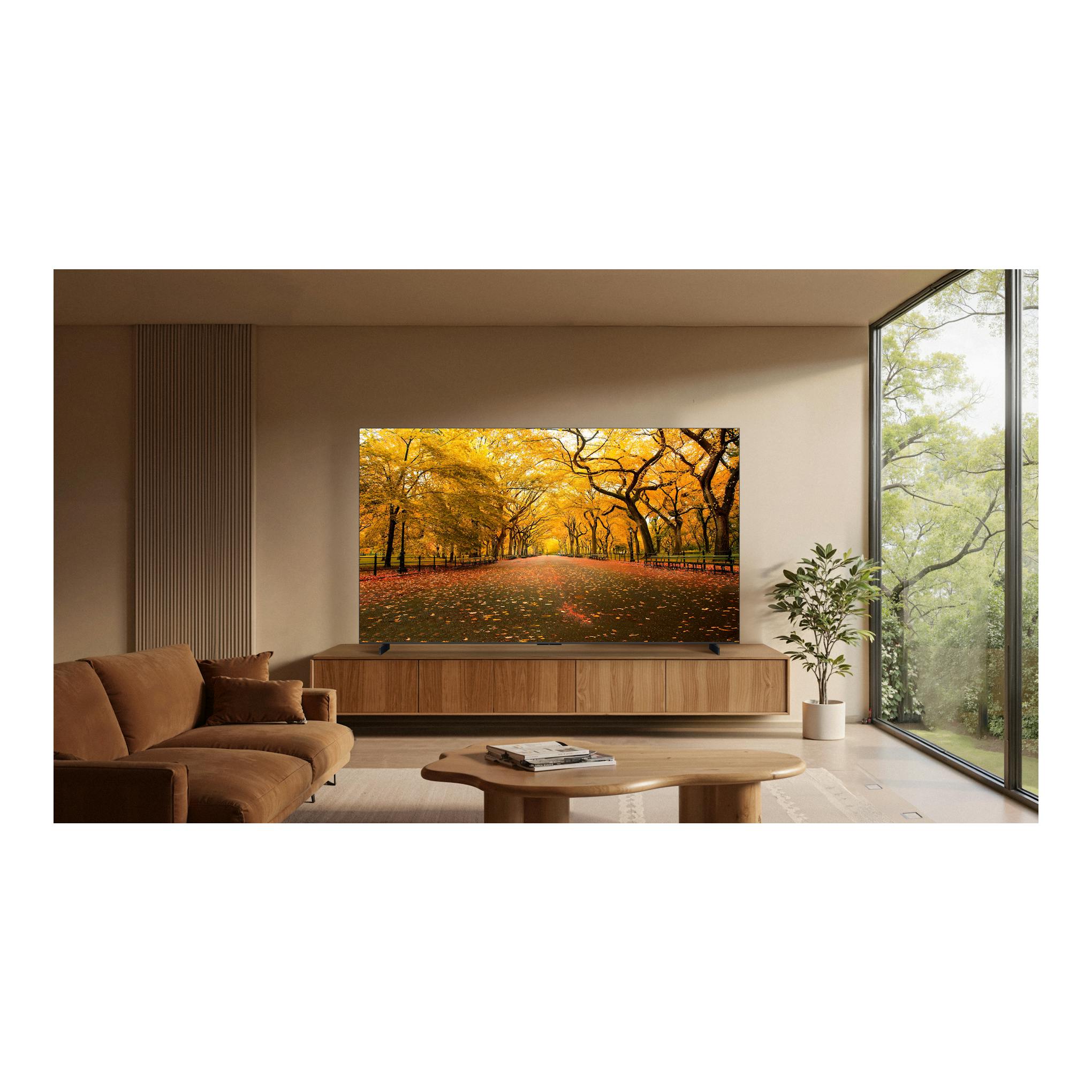 Televisor TCL 98C8L, vista frontal, sobre mueble de madera en salón, con escena de parque otoñal en pantalla.